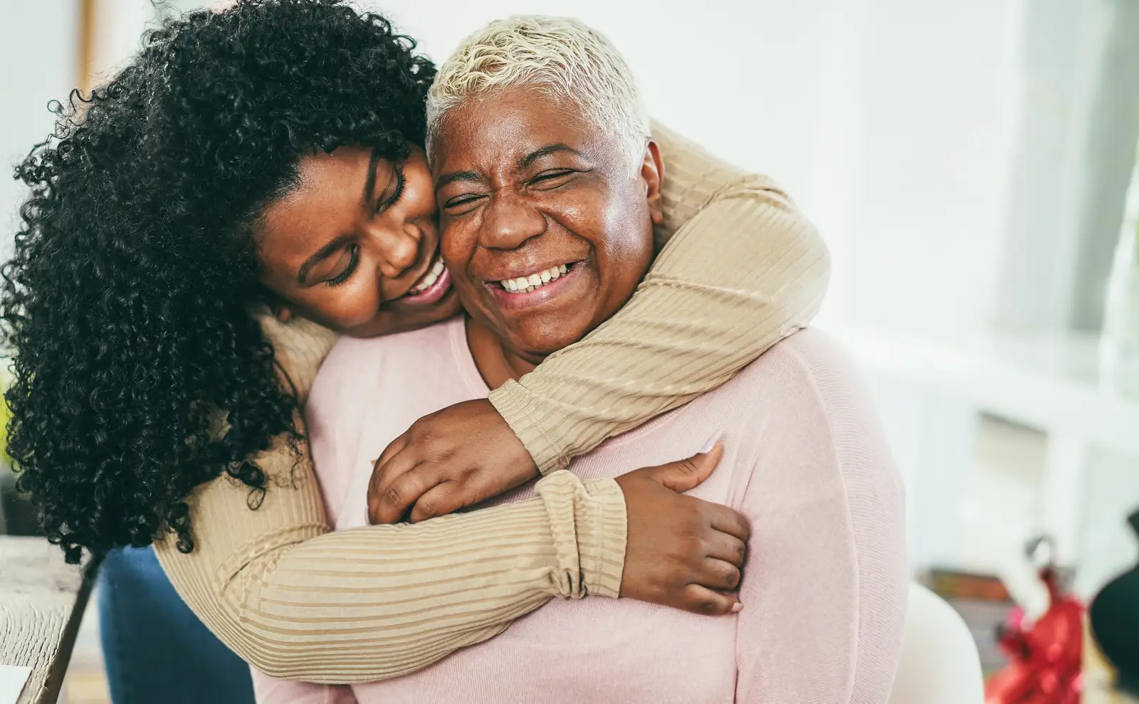A woman with curly hair is smiling and hugging an older woman with white hair in a living room