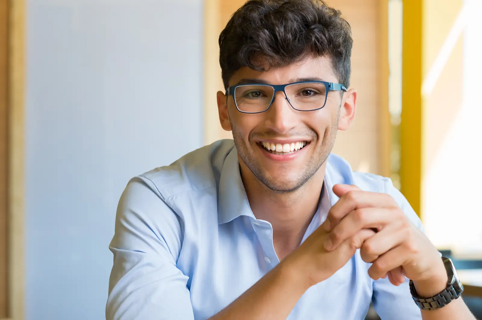 A smiling young man with glasses wearing a light blue button up shirt sits at a table.