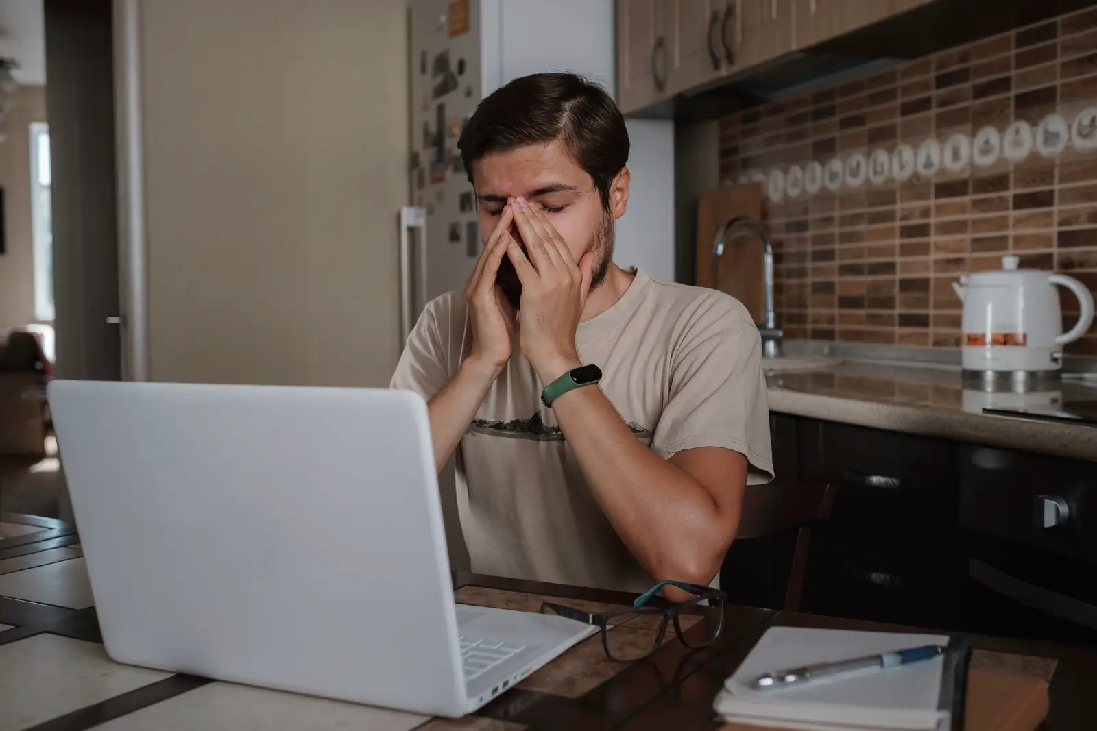 A man sitting at a table in the kitchen with his hands on his face, looking tired and stressed