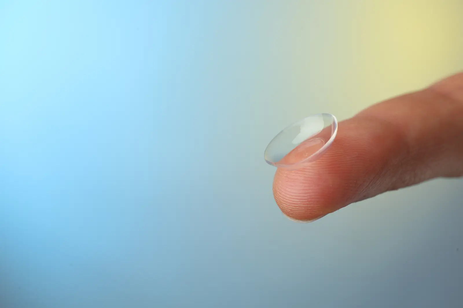 A finger holding a clear contact lens against a blue and yellow background