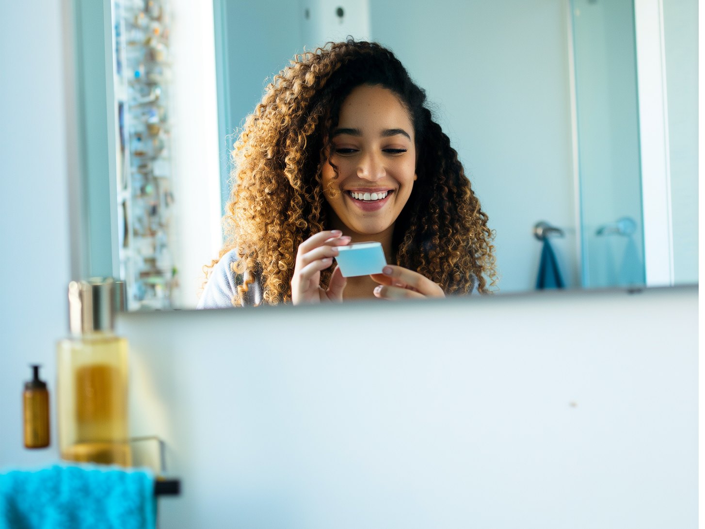 A smiling young woman holding a contact lens case at her bathroom mirror